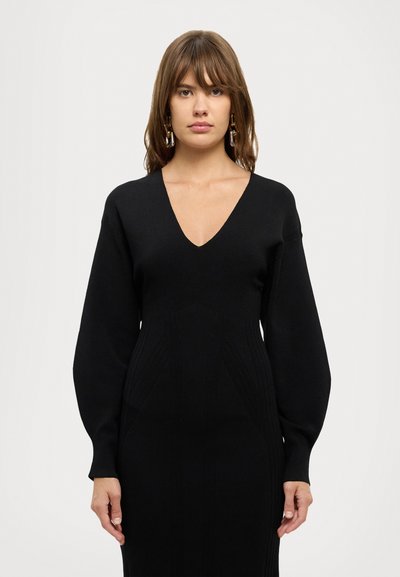 Rochie tricotată neagră cu decolteu în V, bust ajustat și mâneci lungi, largi. Prezintă o textură ribbed și detalii cu cusături verticale.
