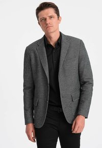 Blazer de espiga gris con solapa de muesca, diseño de un solo botón y dos bolsillos delanteros, sobre una camisa negra y pantalones negros.