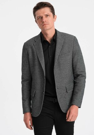 Grau kariertes Blazer mit Reverskragen, einreihiger Ausführung und zwei Vorderaschen, über einem schwarzen Hemd und schwarzen Hosen.