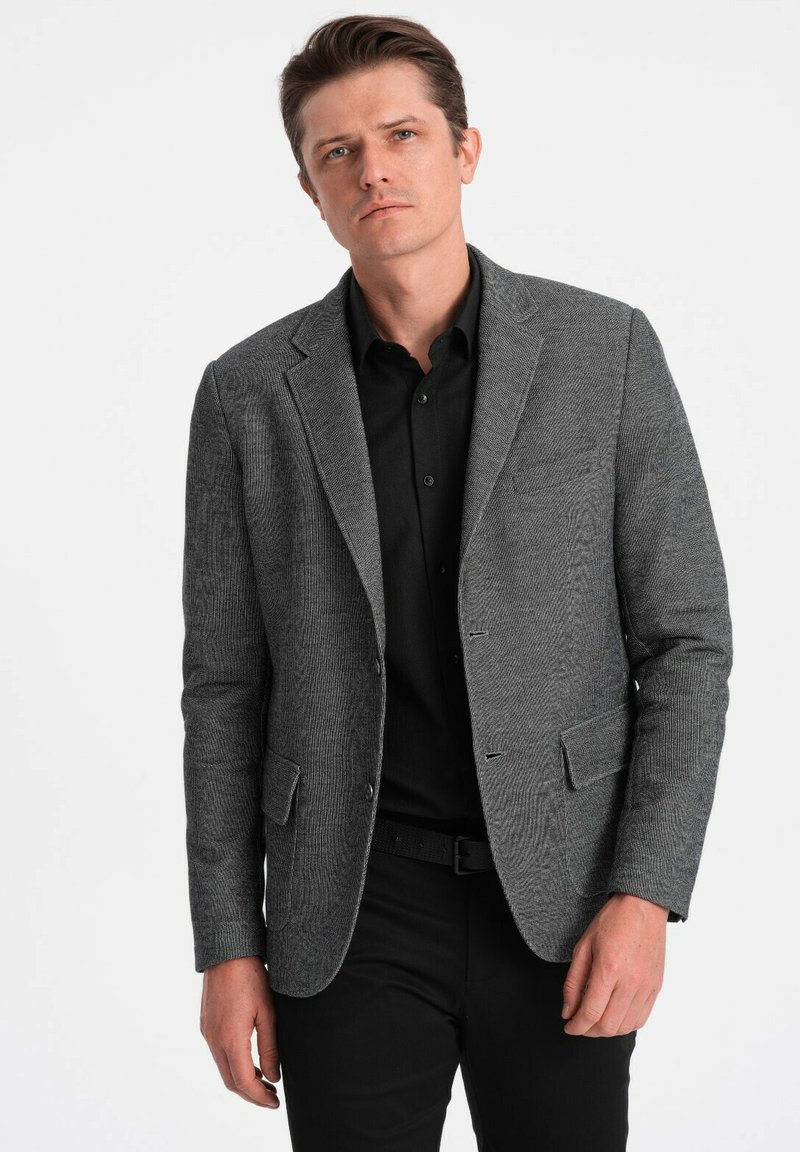 Blazer de espiga gris con solapa de muesca, diseño de un solo botón y dos bolsillos delanteros, sobre una camisa negra y pantalones negros.