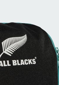 Nahaufnahme einer schwarzen Stofftasche mit einem weißen Farnlogo und dem Teiltext "ALL BLACKS" neben einem türkisfarbenen Reißverschluss und Stoffrand.