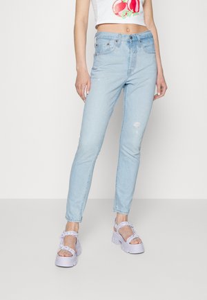 Frau trägt hellblaue High-Waist-Jeans mit leichten Used-Effekten, weiße klobige Sandalen und ein weißes, kurzes Top mit Kirschenmotiv.