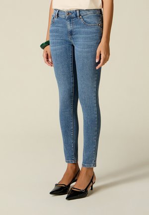 Nainen pukeutunut vaaleansiniin skinny-farkkuihin ja mustiin, kärjestä kapeisiin slingback-korkoihin, seisoo yksivärisen taustan edessä.