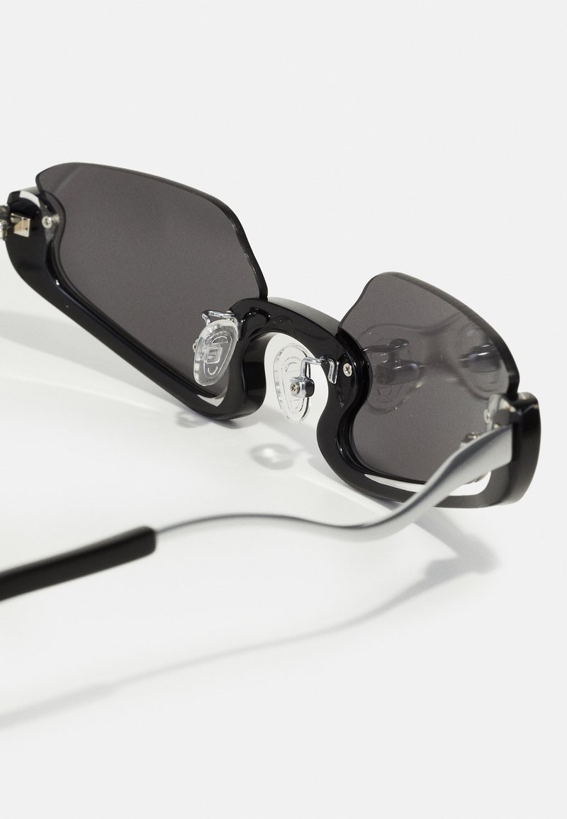 Occhiali da sole neri con grandi lenti grigie opache. Presentano un design della montatura ondulato, con naselli trasparenti e aste sottili e curve.