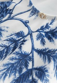 Vitt tyg med ett blått botaniskt tryck som visar detaljerade blad, tillsammans med en gyllene cirkulär clasp under kragen.