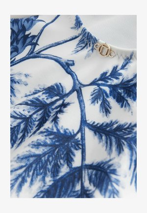 Vitt tyg med ett blått botaniskt tryck som visar detaljerade blad, tillsammans med en gyllene cirkulär clasp under kragen.