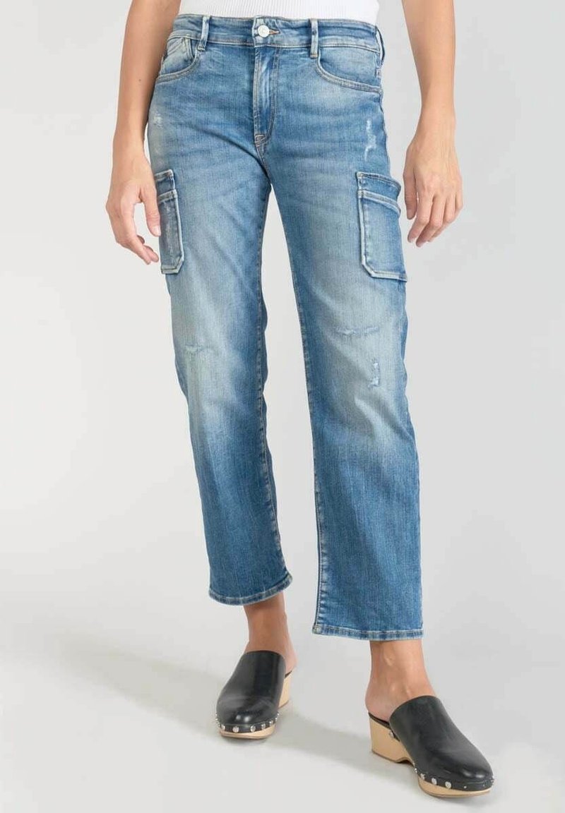 Jeans cargo de mezclilla azul con bolsillos laterales, llevados por una persona de pie, combinados con zuecos de madera negra con tachuelas en los tacones.
