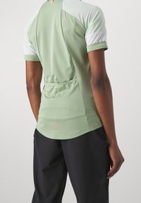 Vaaleanvihreä urheilupaita, jossa on valkoisia verkkokangasaksentteja, lyhyet hihat ja takatasku. Paidassa on pyöristetty helma ja logoyksityiskohta alareunassa.