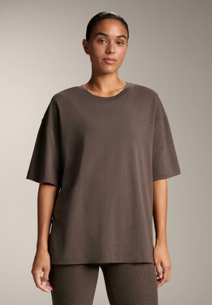 Tricou oversized maro, realizat din bumbac moale. Are manșete scurte, guler rotund și o croială relaxată. Modelul stă în fața unui fundal neutru.