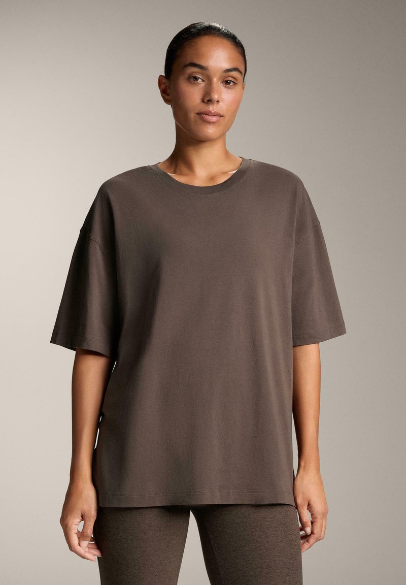 Braunes, oversized T-Shirt aus weicher Baumwolle. Hat kurze Ärmel, einen runden Halsausschnitt und eine lockere Passform. Das Modell steht vor einem neutralen Hintergrund.