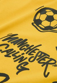 Gul bomulds T-shirt med sort trykt tekst og et fodboldgrafik. Teksten lyder "Manchester Calling" med en fed, legesyg skrifttype.