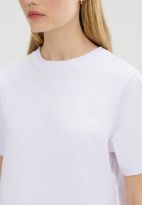 Vrouw met lang blond haar draagt een eenvoudige witte t-shirt met korte mouwen en een subtiel reliëf logo op de borst, tegen een neutrale achtergrond.