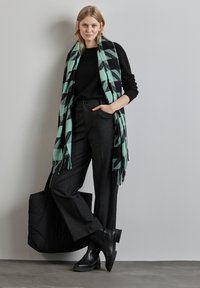 Suéter negro, bufanda a cuadros negra y menta con flecos, pantalones de pierna ancha grises, botas negras hasta el tobillo y un bolso tote acolchado negro.