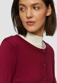 Bordeauxkleurige cardigan met een ronde hals, knoopsluiting en geribbelde randen, gelaagd over een crèmekleurige coltrui. Zachte textuur, getailleerd ontwerp.