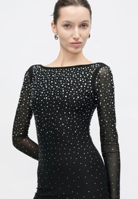 Γυναίκα με μαύρο διαφανές φόρεμα μακρυμάνικο, διακοσμημένο με διάσπαρτες ασημένιες στρας, συνδυασμένο με ασημένια σκουλαρίκια κρίκους και τα μαλλιά της τραβηγμένα πίσω.