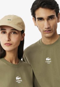 Due persone che indossano camicie coordinate in oliva di Lacoste Paris; una delle due indossa un cappellino beige con il logo del coccodrillo Lacoste.