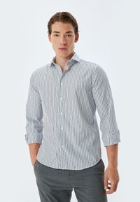 Camisa blanca de cuello con finas rayas verticales azules, de mangas largas remangadas en los puños, y botones de plástico. Combinada con pantalones grises.