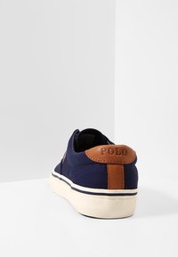 Námornícky plátené tenisky s hnedou koženou patou, bílou gumovou podešví a kontrastním černým proužkem na podešvi. Kulatý tvar, minimalistický design.