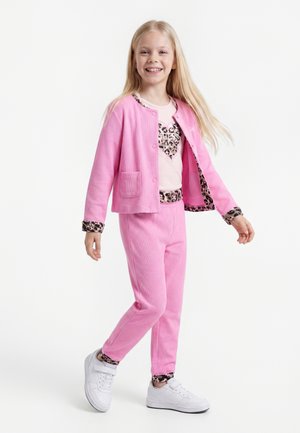 Fille souriante portant une veste et un pantalon roses avec des accents imprimés léopard, baskets blanches, debout devant un fond blanc uni.