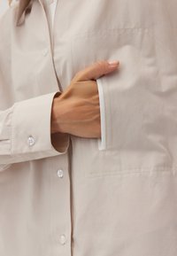 Dłoń częściowo włożona w przednią kieszeń beżowej koszuli z długim rękawem, zapinanej na biały guzik, z widocznym przeszyciem.