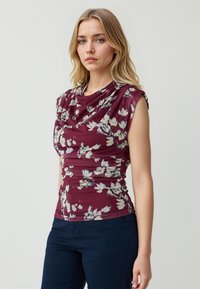 Top floreale con scollatura drappeggiata, dettagli arricciati e maniche a cappuccio. Il motivo presenta fiori bianchi e gialli su uno sfondo borgogna.
