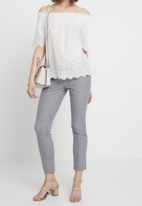 Blusa blanca sin mangas con detalles de ojetes y dobladillo ondulado, combinada con pantalones ajustados de cuadros en blanco y negro. Sandalias de tacón bloque en color beige.