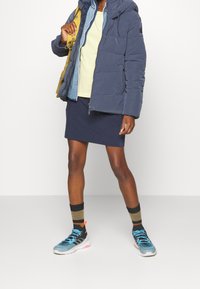 Navy puffer jacket with a hood, beige shirt, navy skirt, and multicolored sneakers. Features elastic socks with horizontal stripes.