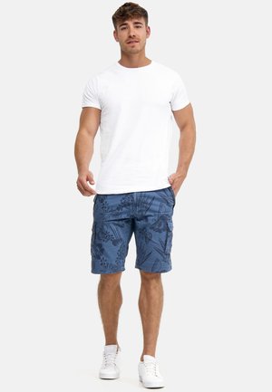 Junger Mann stehend, trägt ein weißes T-Shirt, blaue florale Cargoshorts und weiße Sneakers vor einem einfarbigen hellen Hintergrund.