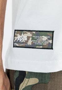 Tricou alb cu mânecă scurtă, cu un eticheta rectangulară camuflată, având cusături negre și text alb. Materialul pare moale și ușor.