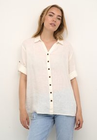 Linned knap-op skjorte i off-white med v-hals, korte rullede ærmer og brune knapdetaljer, parret med lyseblå slidte jeans.