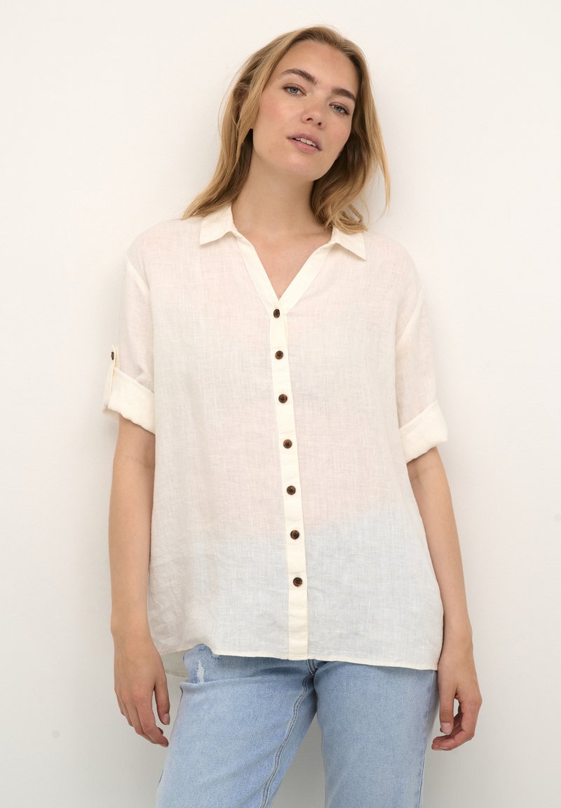 Linnen button-up shirt in gebroken wit met een v-hals, korte opgerolde mouwen en bruine knoopdetails, gecombineerd met lichtblauwe distressed jeans.