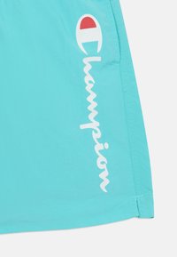 Pantaloni corti blu acqua realizzati in un tessuto leggero, con logo "Champion" bianco e un accento rosso in un design circolare.
