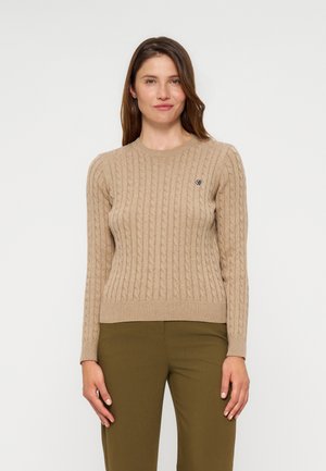 Frau trägt einen beigen, kabelgestrickten Pullover mit langen Ärmeln und olivgrüne Hosen, steht vor einem schlichten hellgrauen Hintergrund.