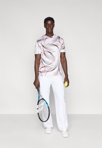 Ενδυμασία για τένις που περιλαμβάνει ένα λευκό πουκάμισο με κόκκινα και μαύρα αφηρημένα σχέδια, συνοδευόμενο από λευκό παντελόνι, κρατώντας ρακέτα και μπάλα τένις.