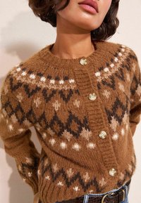 Naisel on seljas pruun mustriga kudumkardigan koos kreemiste nööpidega, mille all on sinised teksad ja tume vöö.