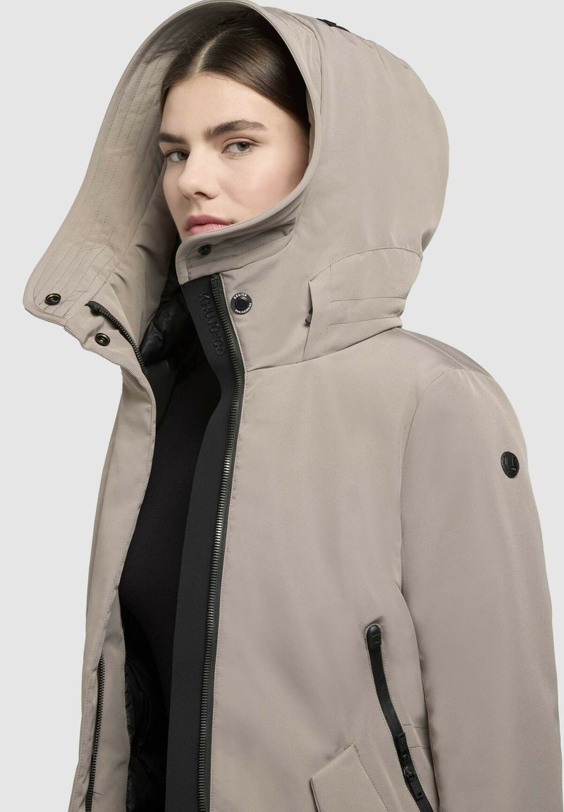 Giacca impermeabile beige con un ampio cappuccio, dettagli neri, zip frontale e tasche laterali; texture liscia e caratteristiche regolabili.