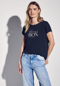 T-shirt noir à manches courtes avec "BON" imprimé en crème, associé à un jean en denim bleu clair. Tissu lisse, coupe décontractée et style casual.