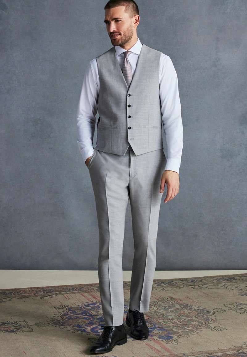 Costume gris ajusté avec un gilet cintré à fermeture à cinq boutons, associé à une chemise blanche et des chaussures noires. Pantalons à pli avant.