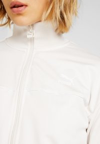 Giacca bianca con zip e colletto alto, texture a coste e sottili cuciture orizzontali. Presenta un piccolo logo Puma bianco sul petto.