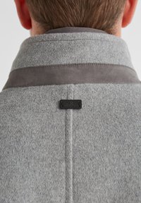 Graue Wolle-Mischjacke mit einem glatten grauen Kragen, ausgestattet mit einem dezenten Logolabel und strukturiertem Stoff. Sichtbare Nähte entlang der Nähte.