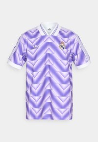 Camiseta morada con un patrón geométrico, con un cuello y puños blancos, logo de Adidas y escudo del Real Madrid en el pecho.