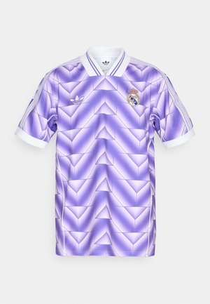 Maglia viola con un motivo geometrico, con colletto e polsini bianchi, logo Adidas e stemma del Real Madrid sul petto.