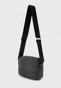 Saco cruzado de tecido cinza com formato retangular, apresentando uma superfície texturizada, alça preta com logótipo e detalhes em hardware hexagonal.