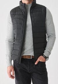 Black, padded vest with a high collar, zip front, and horizontal stitching. Worn over a light gray sweater, paired with dark jeans and a brown belt.