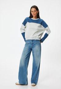 Felpa bloccata nei colori grigio chiaro e blu, con maniche lunghe, scollatura rotonda e vestibilità rilassata, abbinata a jeans blu a gamba larga e sneakers.