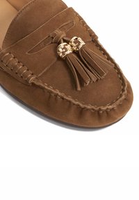 Brown suede moccasin shoe with tassel accents and gold hardware, featuring stitched detailing and a rounded toe design.