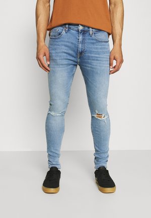 Homme portant un jean slim bleu clair déchiré et des baskets noires avec des semelles en gomme, debout devant un fond clair uni.