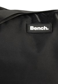 Zwarte stoffen tas met een gestructureerd oppervlak, voorzien van een rechthoekig zwart label met "Bench." in witte letters.