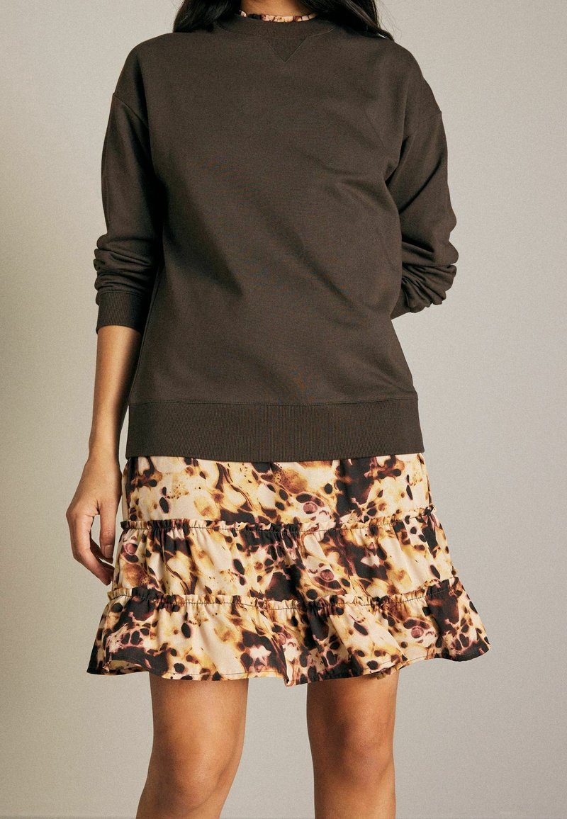Sweatshirt marron avec des poignets et un ourlet côtelés, associé à une jupe à volants florale dans des tons beiges et marron, présentant un design superposé.