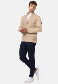 Suéter beige con cremallera y patrón de punto texturizado, camisa blanca de cuello contrastante debajo, combinado con pantalones azul marino y zapatillas ligeras.